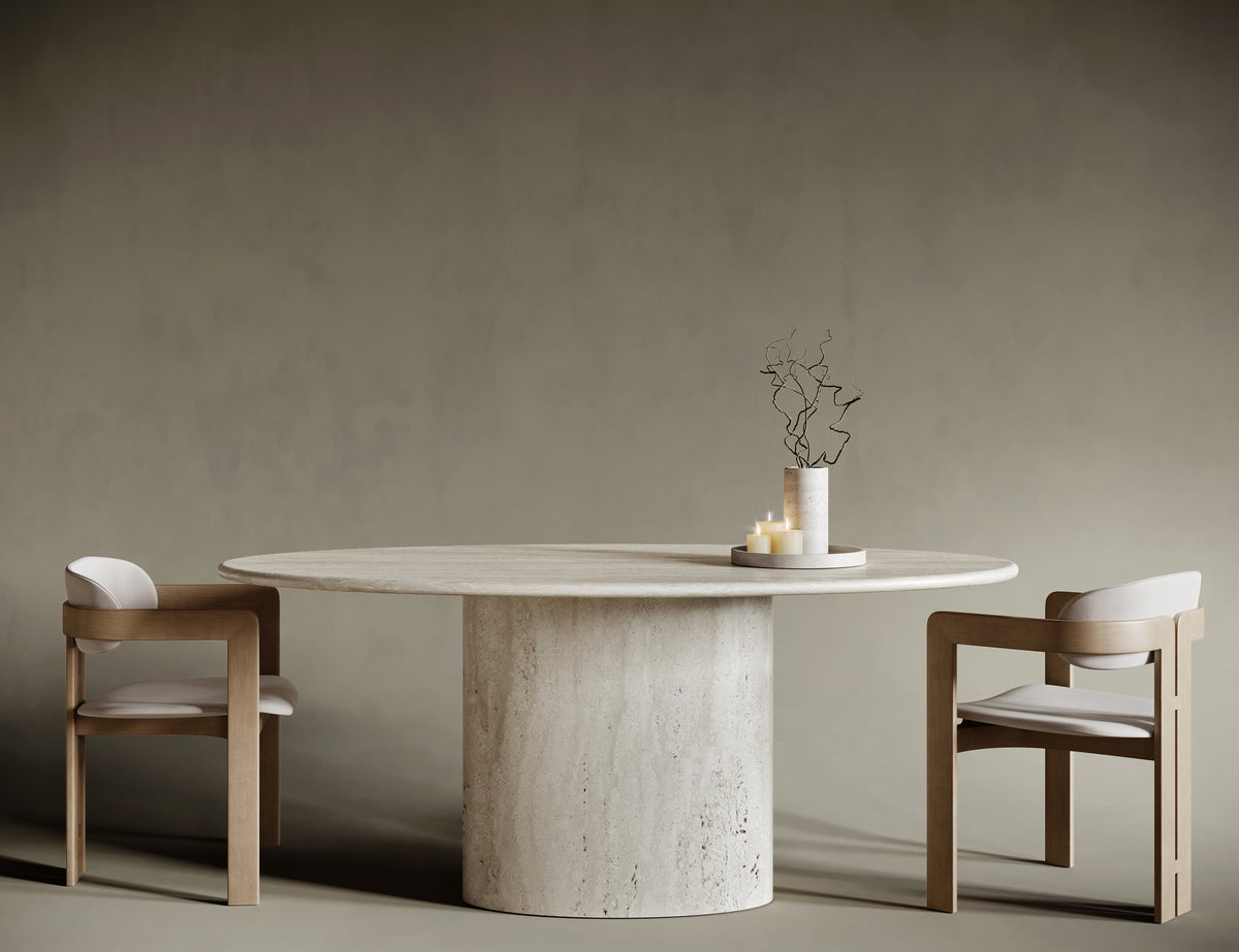 Minimalist dining room featuring a round ellipse travertine dining table with a cylindrical pedestal base, paired with two modern wooden chairs upholstered in cream fabric. The natural stone table showcases a textured surface, complemented by a simple centerpiece of twisted branches and candles. Set against a muted taupe background, this elegant interior highlights contemporary furniture design and natural materials