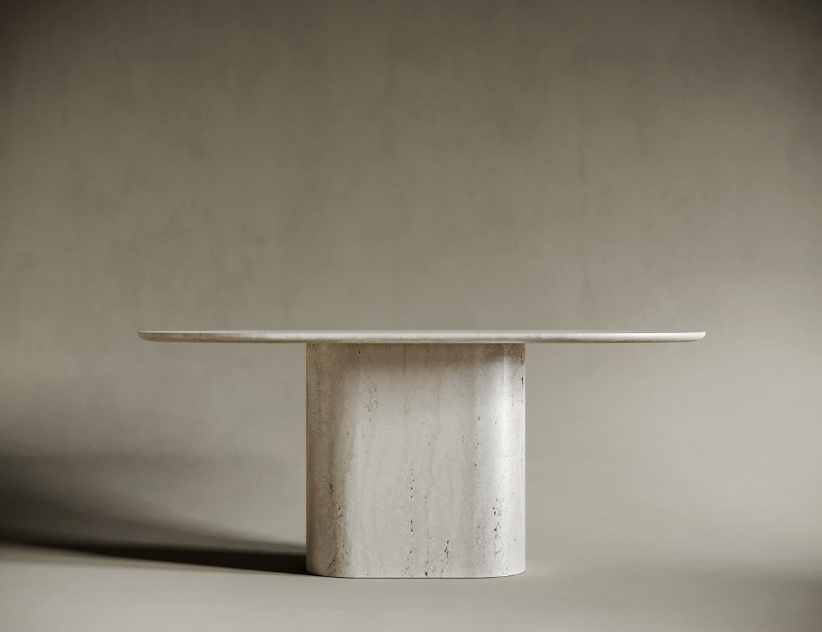 Elegant dining space centered around an oval travertine table with a monolithic pedestal base. The natural stone surface, paired with light wood chairs and cream upholstery, creates a harmonious blend of organic materials and modern design. The muted gray-green backdrop enhances the table's sculptural presence, making it a focal point of refined interiors.