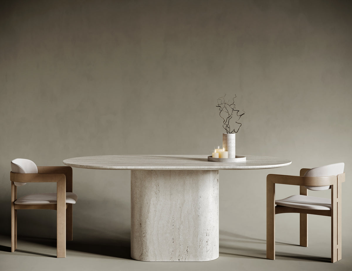 Minimalist dining room featuring an oval travertine dining table with a sculptural single pedestal base. Paired with two modern wooden chairs upholstered in light beige fabric, the table showcases natural stone textures and timeless design. Set against a muted olive-beige background, this serene interior highlights luxury craftsmanship and contemporary elegance.