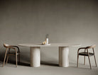 Elegant oval travertine dining table supported by a single cylindrical leg, combining fluid curved lines with the solidity of natural stone; ideal for sophisticated dining rooms where form meets function with architectural clarity