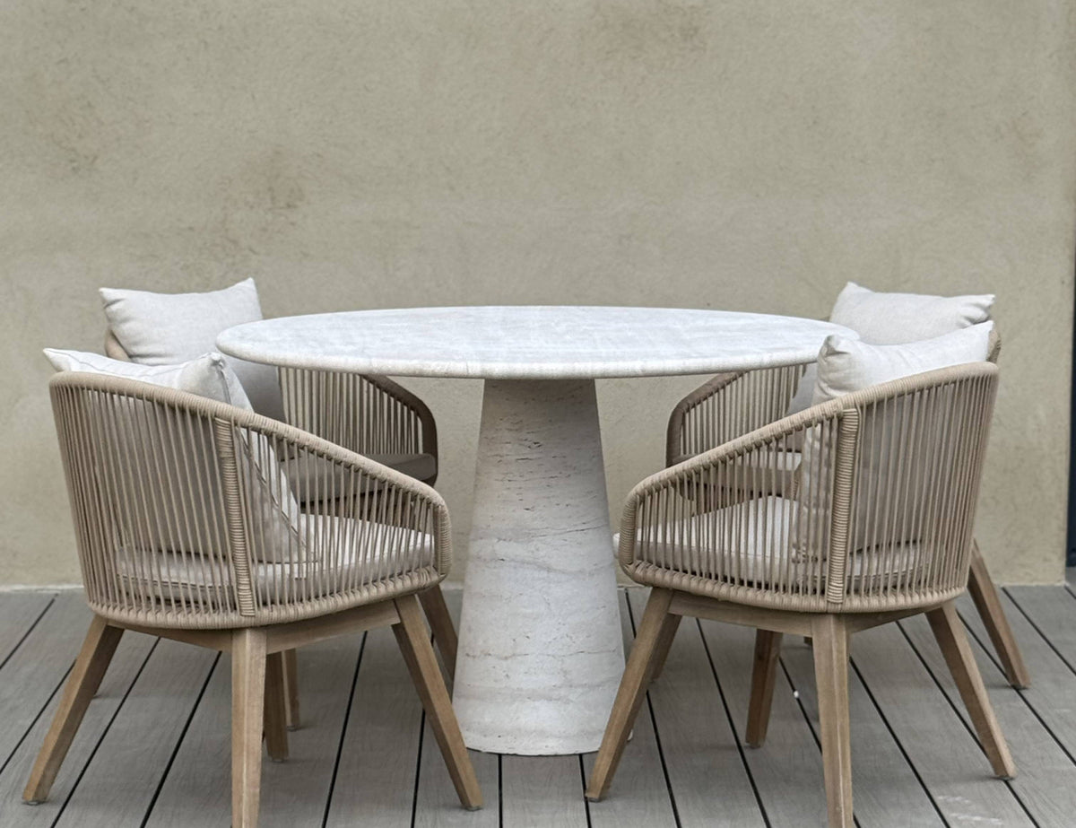 Round outdoor travertine table with four matching chairs on a wooden deck against a neutral wall.
