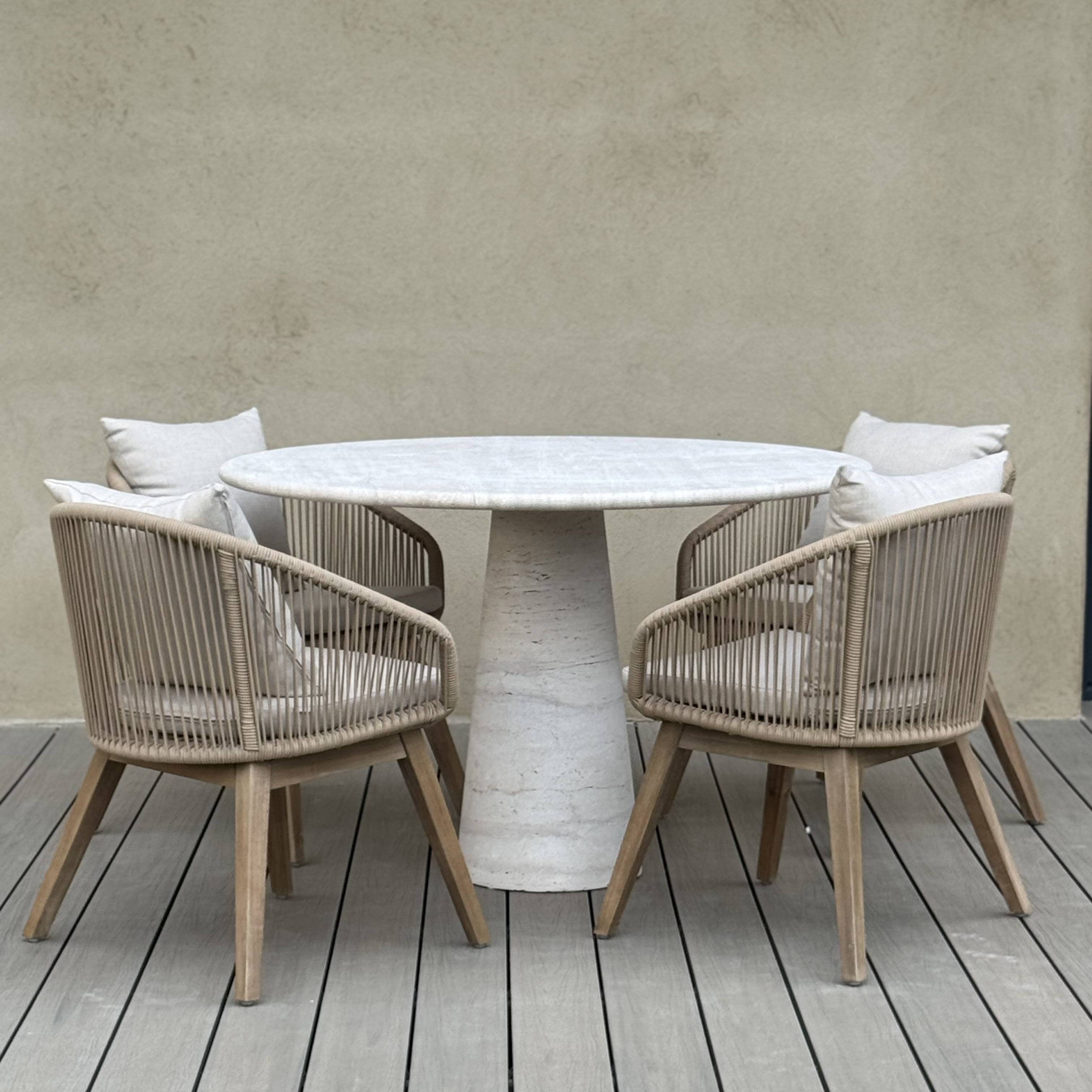 Round outdoor travertine table with four matching chairs on a wooden deck against a neutral wall.