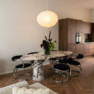 A modern dining room scene featuring an oval marble-top dining table with a sculptural marble pedestal base, surrounded by six black upholstered chairs with chrome curved metal frames. A glowing white paper lantern pendant light hangs above, and a vase of flowers sits on the table.