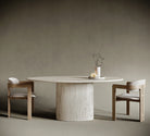 Minimalist dining room featuring a round ellipse travertine dining table with a cylindrical pedestal base, paired with two modern wooden chairs upholstered in cream fabric. The natural stone table showcases a textured surface, complemented by a simple centerpiece of twisted branches and candles. Set against a muted taupe background, this elegant interior highlights contemporary furniture design and natural materials