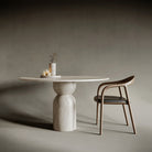 Statement round travertine dining table with an hourglass-shaped base, embodying fluid geometry and balanced tension; crafted from richly textured stone, this piece merges sculptural form with functional design