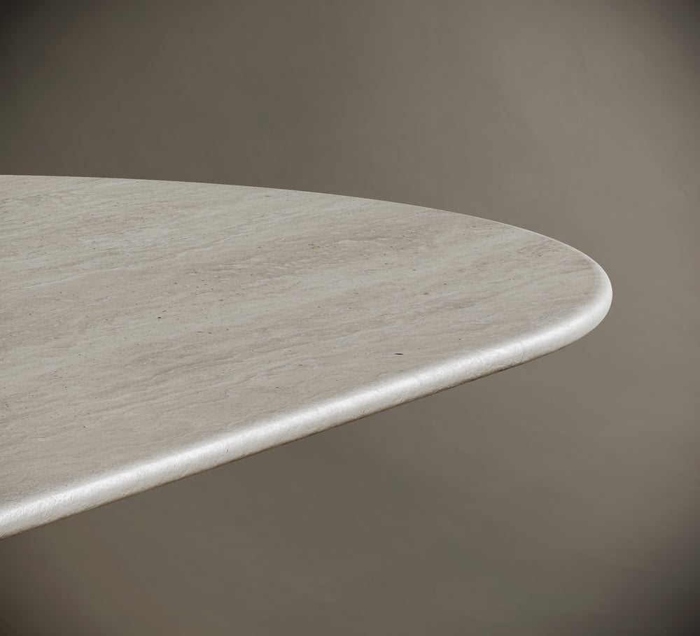 Oval travertine dining table adorned with a matching stone vase holding dried branches and unlit pillar candles. The minimalist decor enhances the table's organic beauty and sculptural presence, perfect for modern interiors