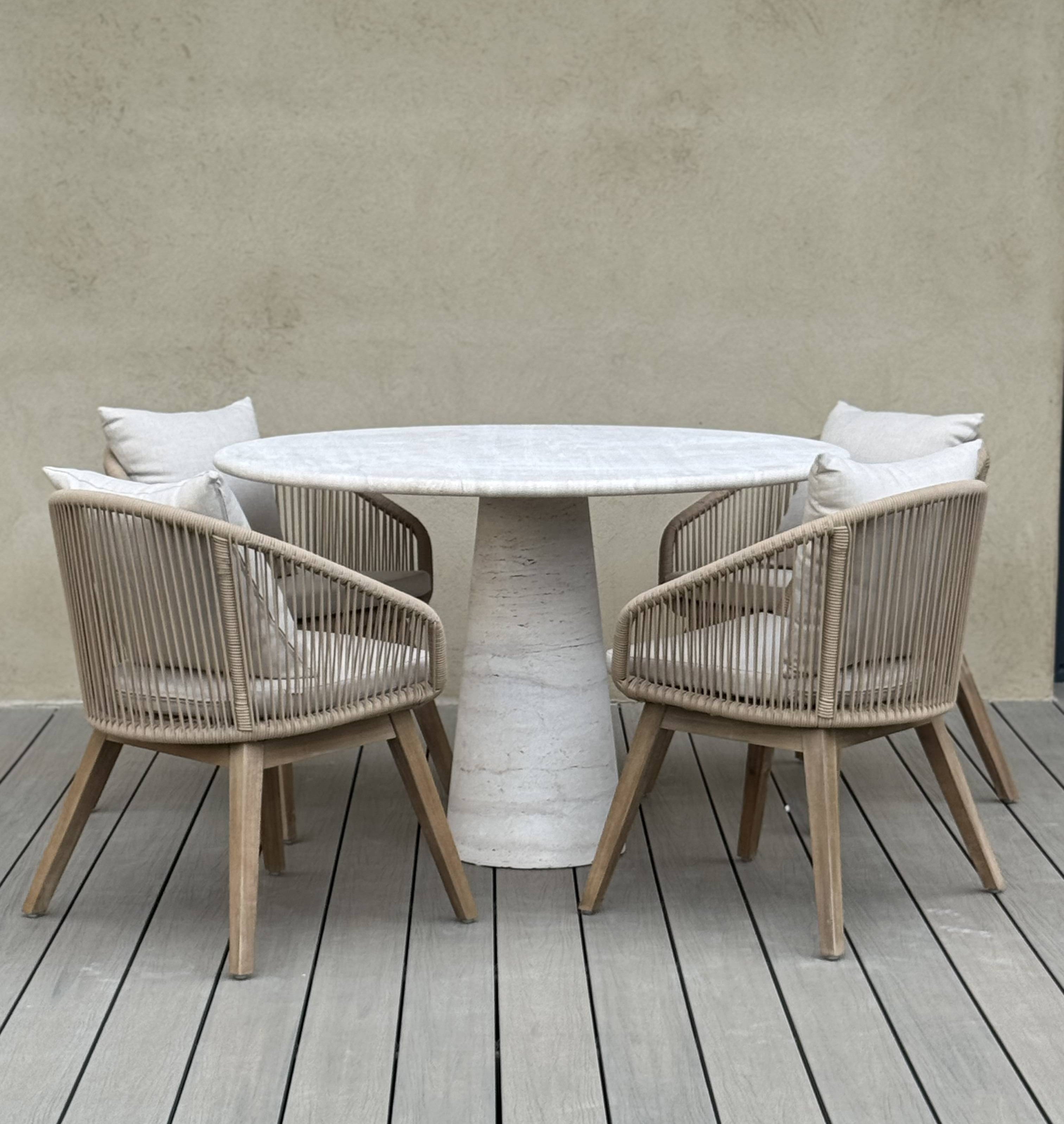 Round outdoor travertine table with four matching chairs on a wooden deck against a neutral wall.