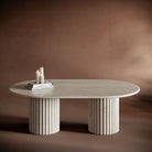 SERENA travertine coffee table with an oval top and fluted cylindrical bases, showcasing natural stone textures.
